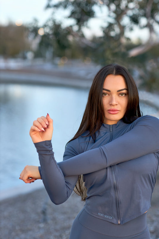 Woman in a gray athletic outfit stretching outdoors by a body of water.